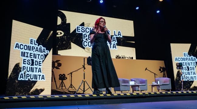 Mujer con gafas hablando por micrófono en un escenario con pantallas gráficas de fondo.