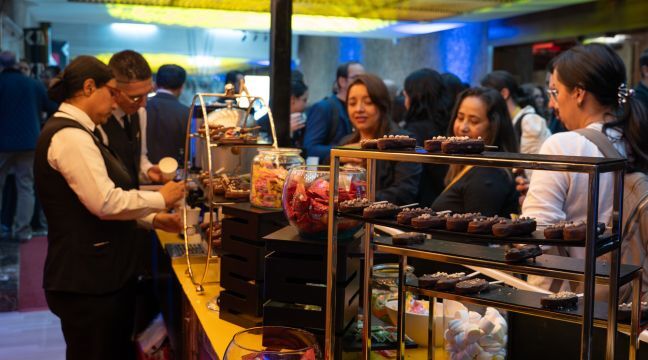 Mesa de refrigerios con postres de chocolate y personas siendo atendidas durante un evento.